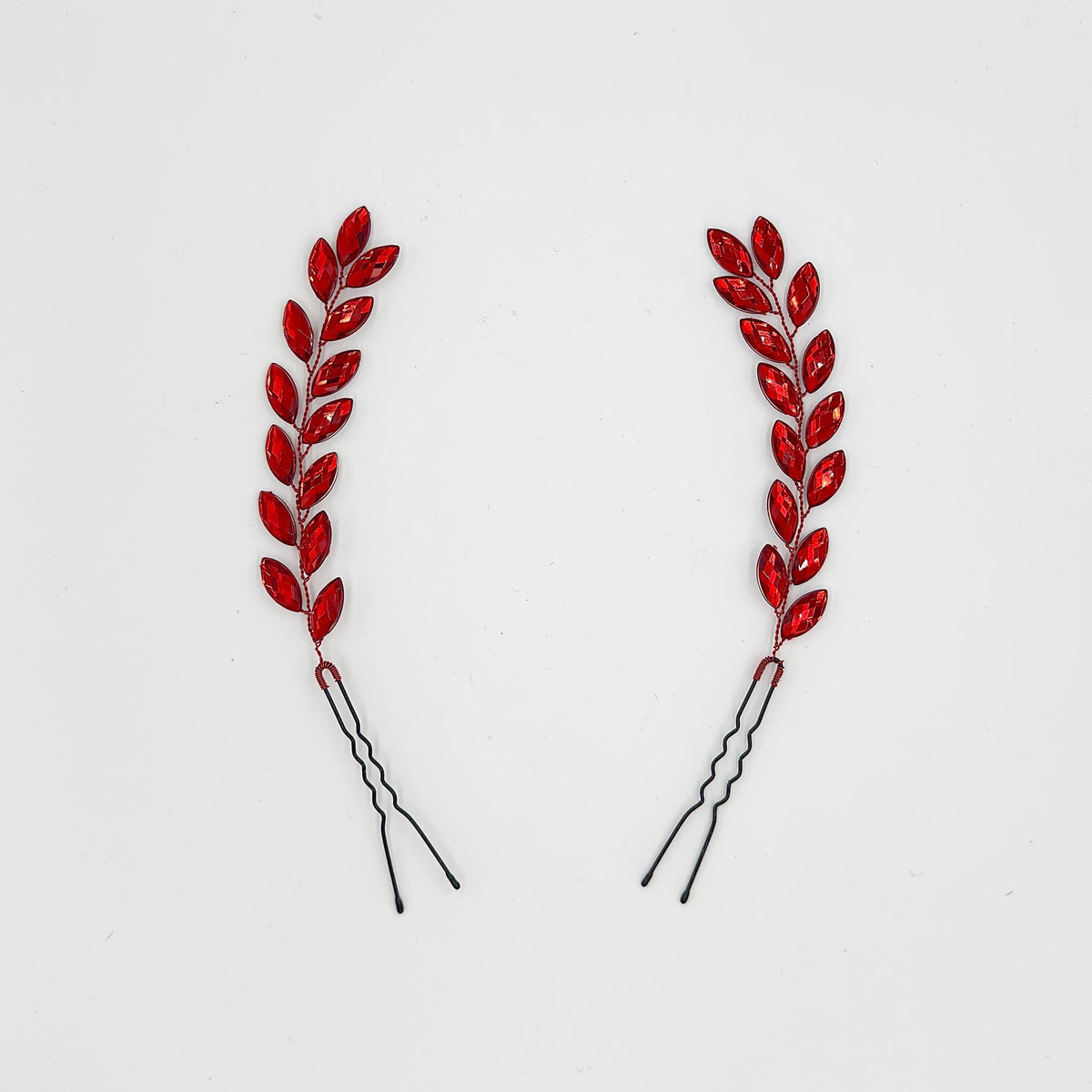 Two long bridal hair pins on a white background with red faceted leaf elements, red branches and black wavy pin bases.