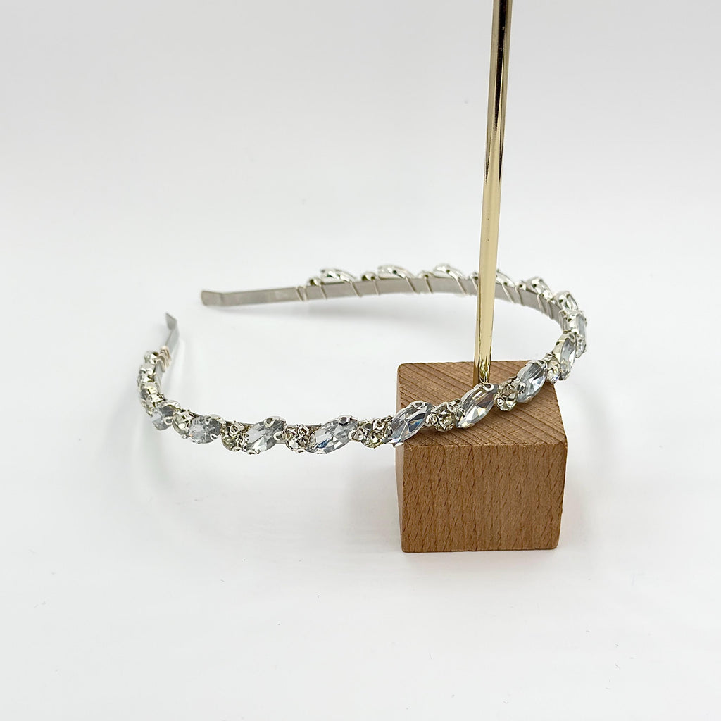 Silver-tone bridal headband on a white background, displayed at an angle over a wooden cube with alternating clear elongated teardrop-shaped and round set stones.