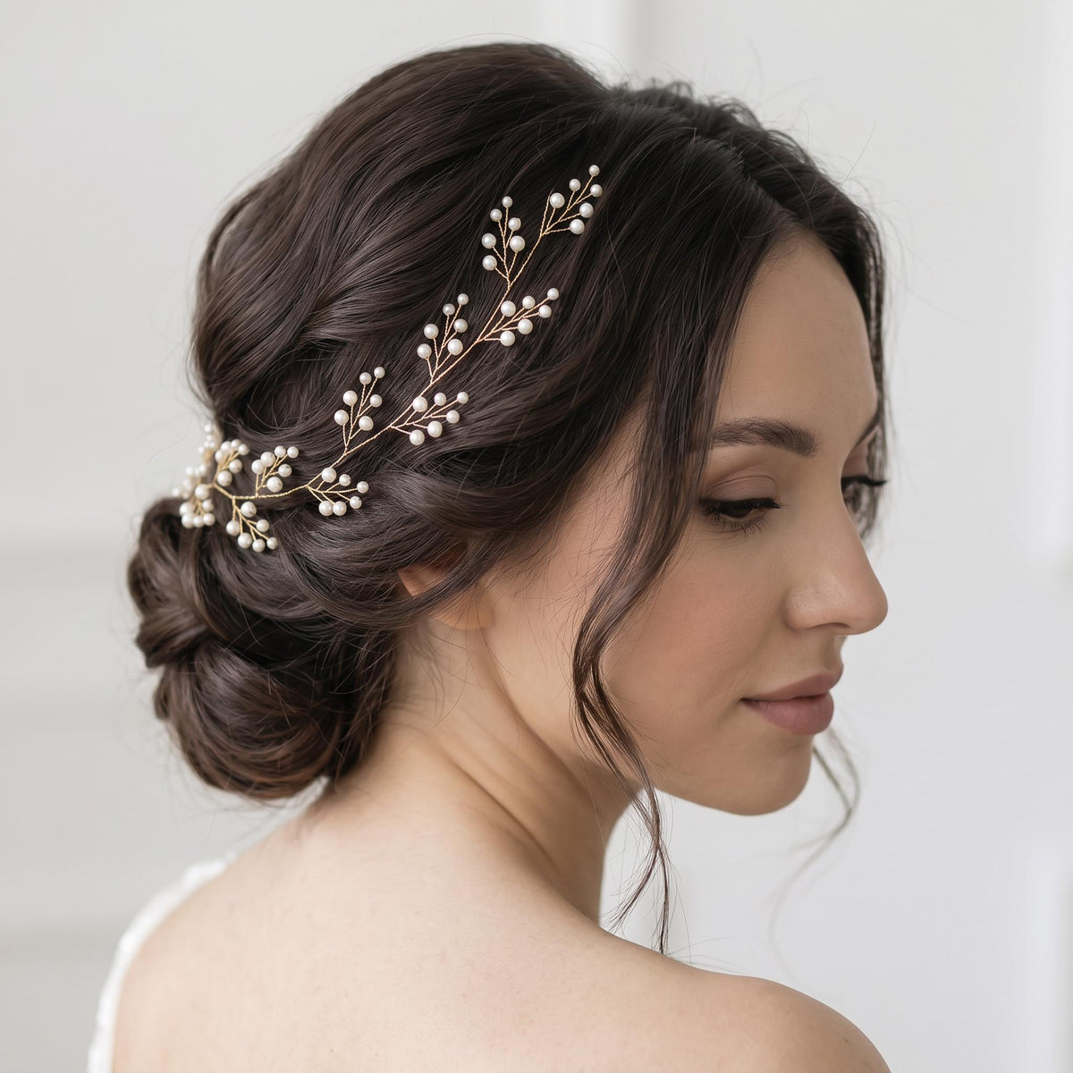 Braut mit seitlich gescheiteltem Haar und tiefem Dutt trägt eine silberfarbene Perlen-Haarranke, deren verzweigte Form sanft vom Vorderkopf zur Hochsteckfrisur verläuft.