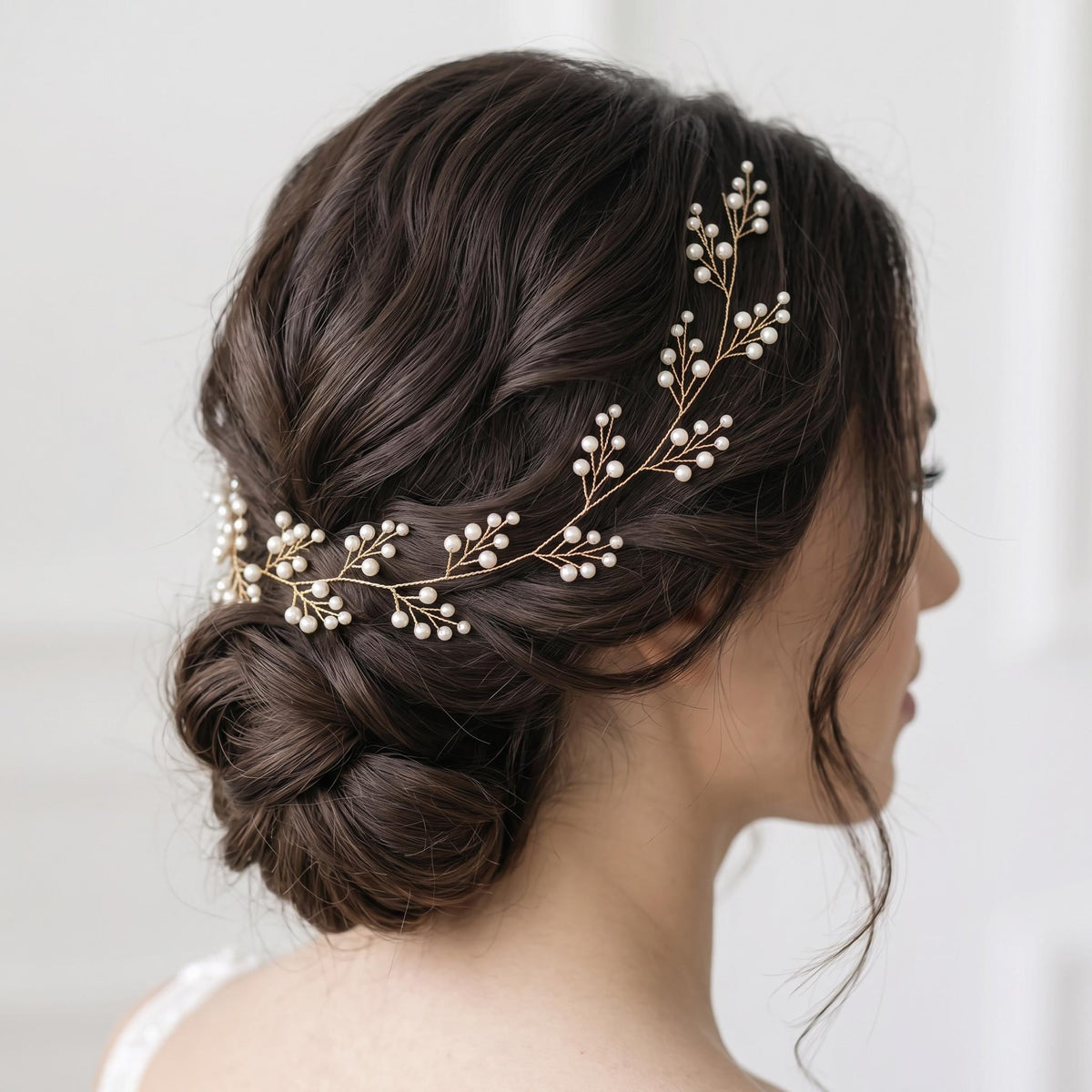 Dark-haired bride wearing a gold-tone flexible pearl hair vine along the side of her head, extending from the front hairline toward a softly styled low bun.