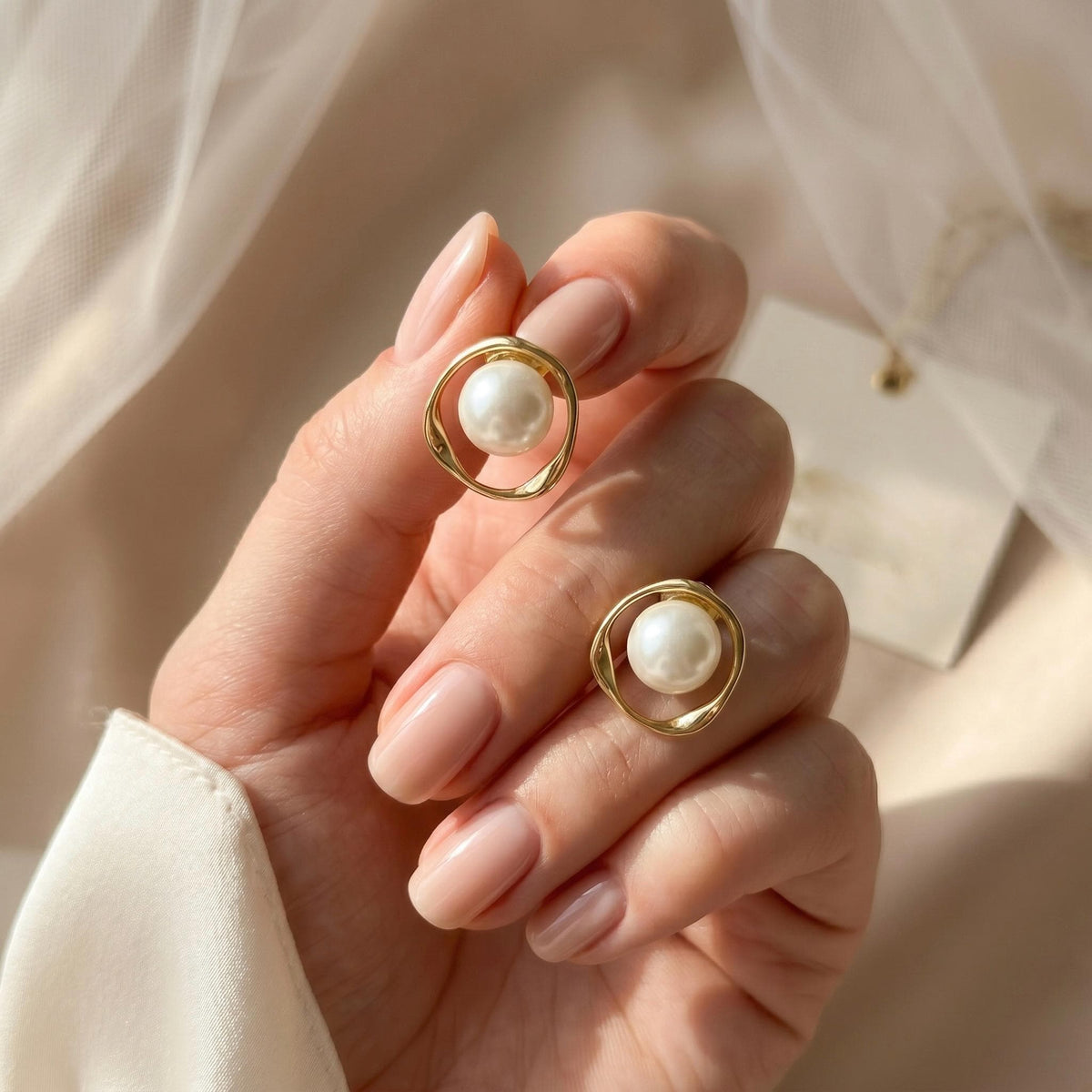 A hand holds two gold-toned stud earrings, each featuring a white pearl centered within a twisted, open circle design.