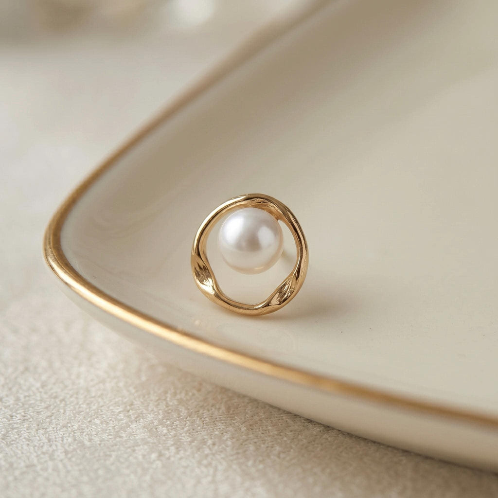 A single gold-toned stud earring, with a white pearl centered in a twisted open circle, rests on a cream-colored dish.