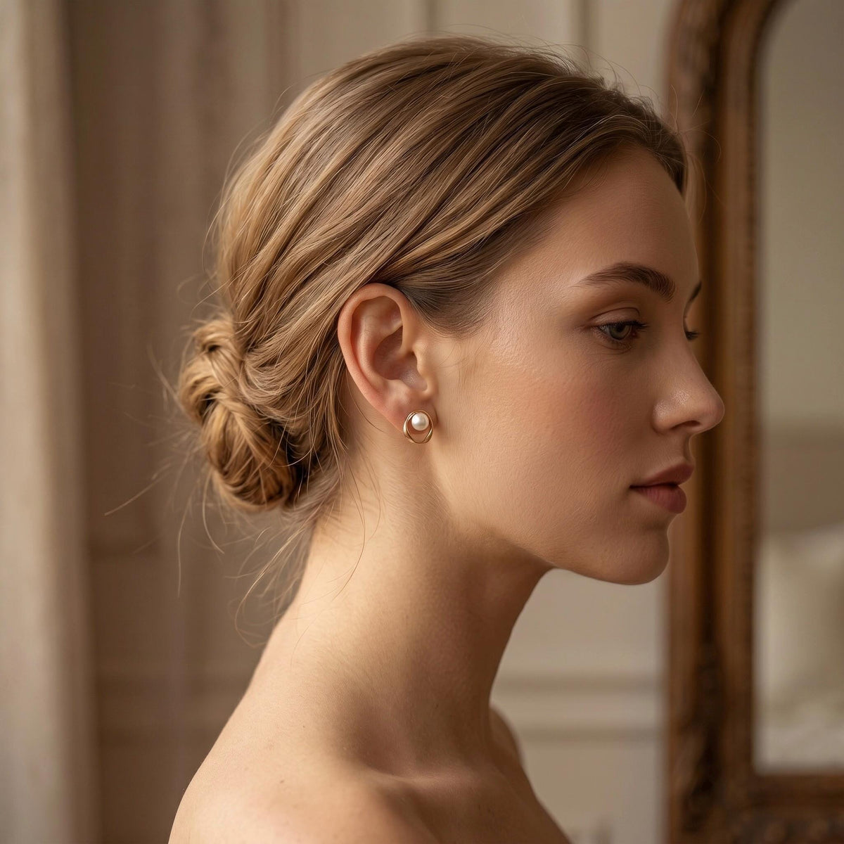 A woman with light brown hair in a bun looks to the side, wearing a gold-toned stud earring with a white pearl.