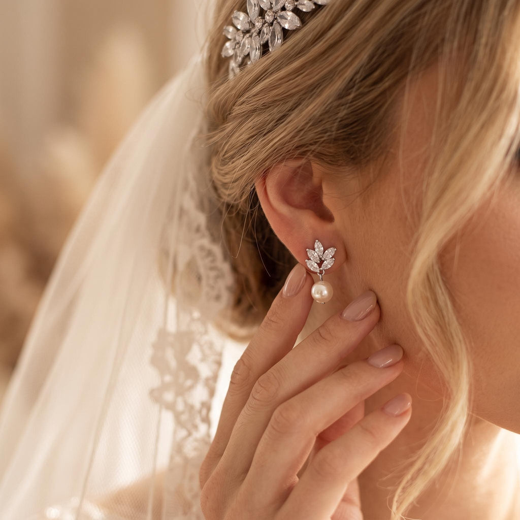 Blonde Braut mit Schleier trägt einen kurzen silberfarbenen Ohrhänger mit einem Zweig aus klaren Marquise-Steinen und einer einzelnen weißen Perle; warmes Seitenlicht betont den Glanz.