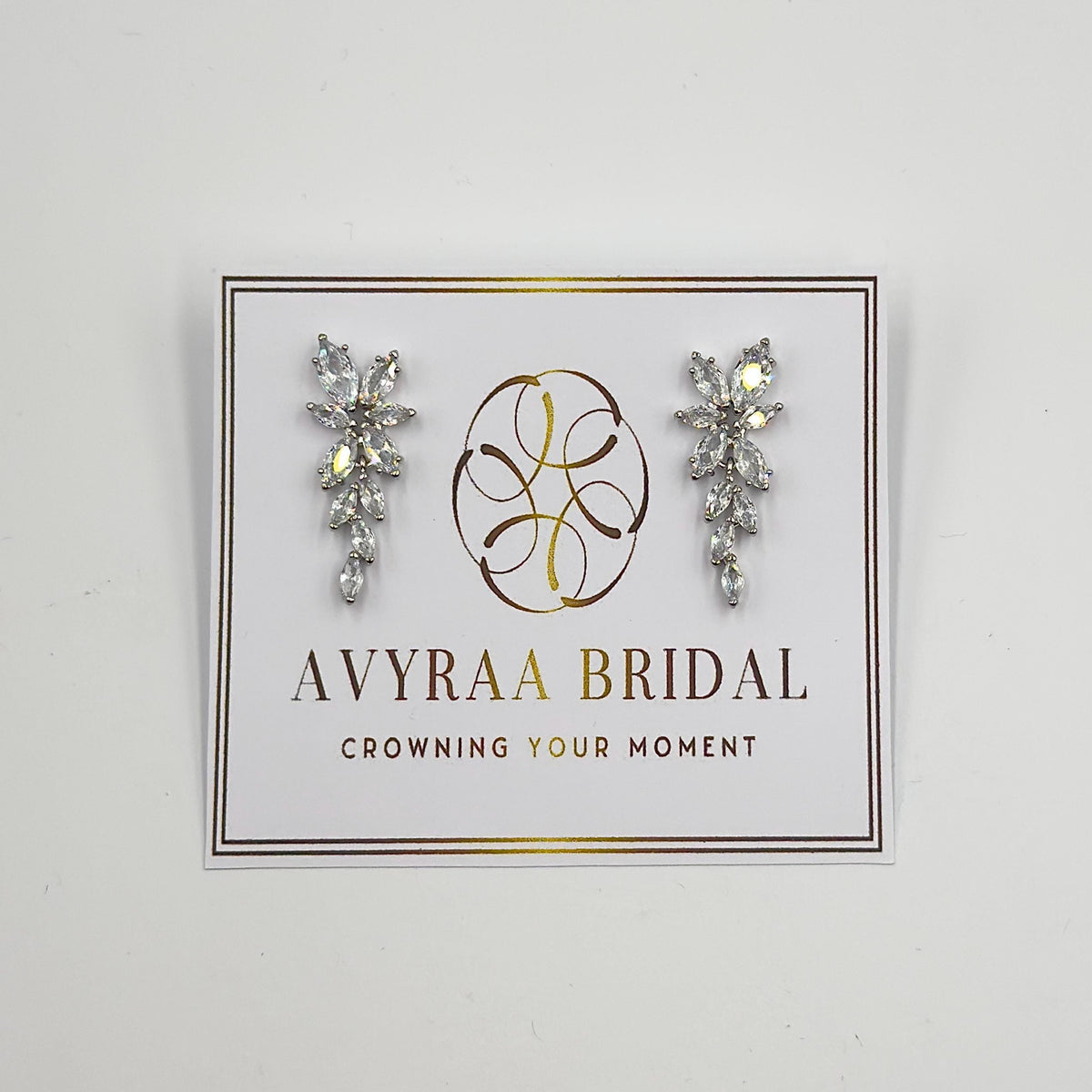 Two silver-toned earrings with clear, marquise-cut gemstones in a floral and leaf-like drop design are displayed on a white card with "AVYRAA BRIDAL" and "CROWNING YOUR MOMENT" printed in black.
