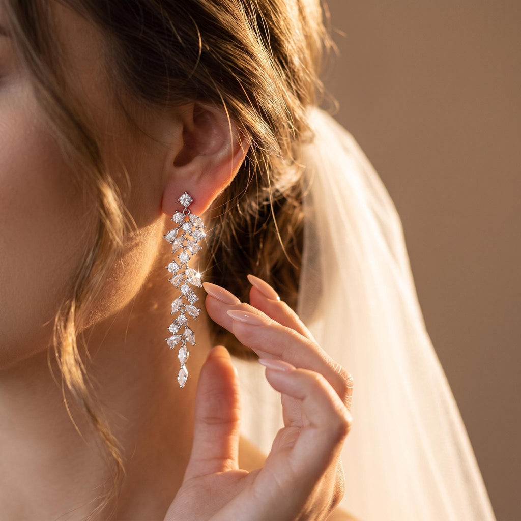 A close-up of a woman's right ear, wearing a long, silver-toned earring with clear, leaf-shaped and round gemstones, her hand gently touching her jawline.