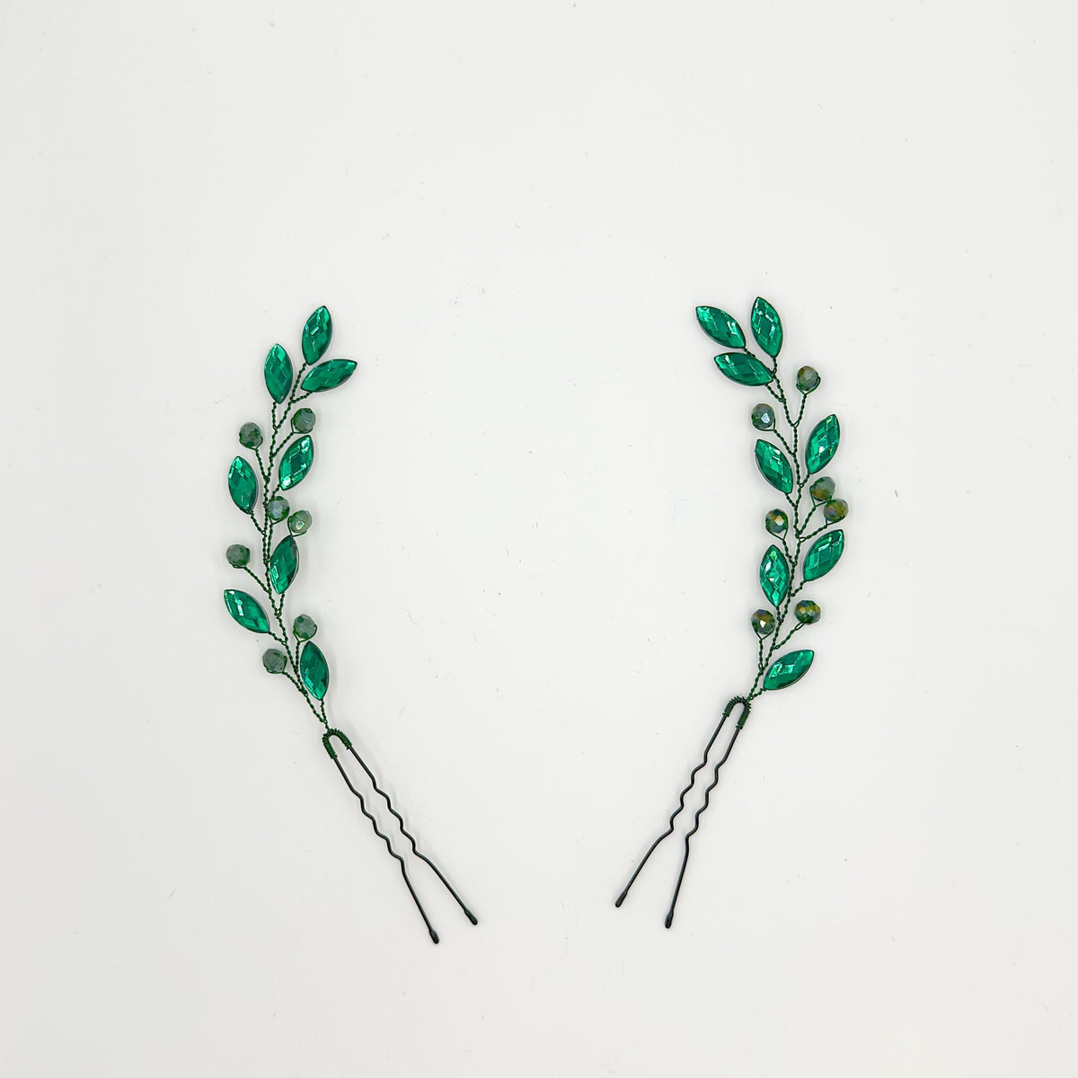 Two green bridal hair pins on a white background with faceted leaf stones, smaller round crystals, dark green branches and black pin bases.