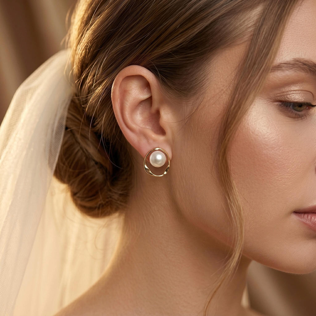 A woman wearing a white veil has a gold-toned stud earring with a white pearl inside a twisted open circle on her earlobe.