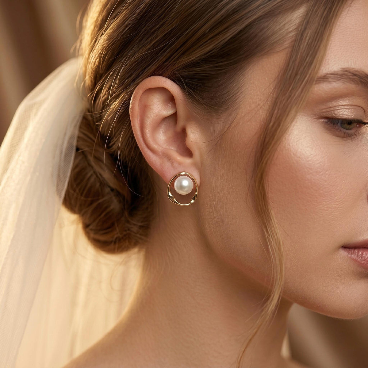 A woman wearing a white veil has a gold-toned stud earring with a white pearl inside a twisted open circle on her earlobe.